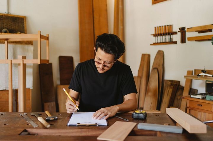 Person in a workshop sketching a design on paper at a wooden workbench, surrounded by wood pieces and tools