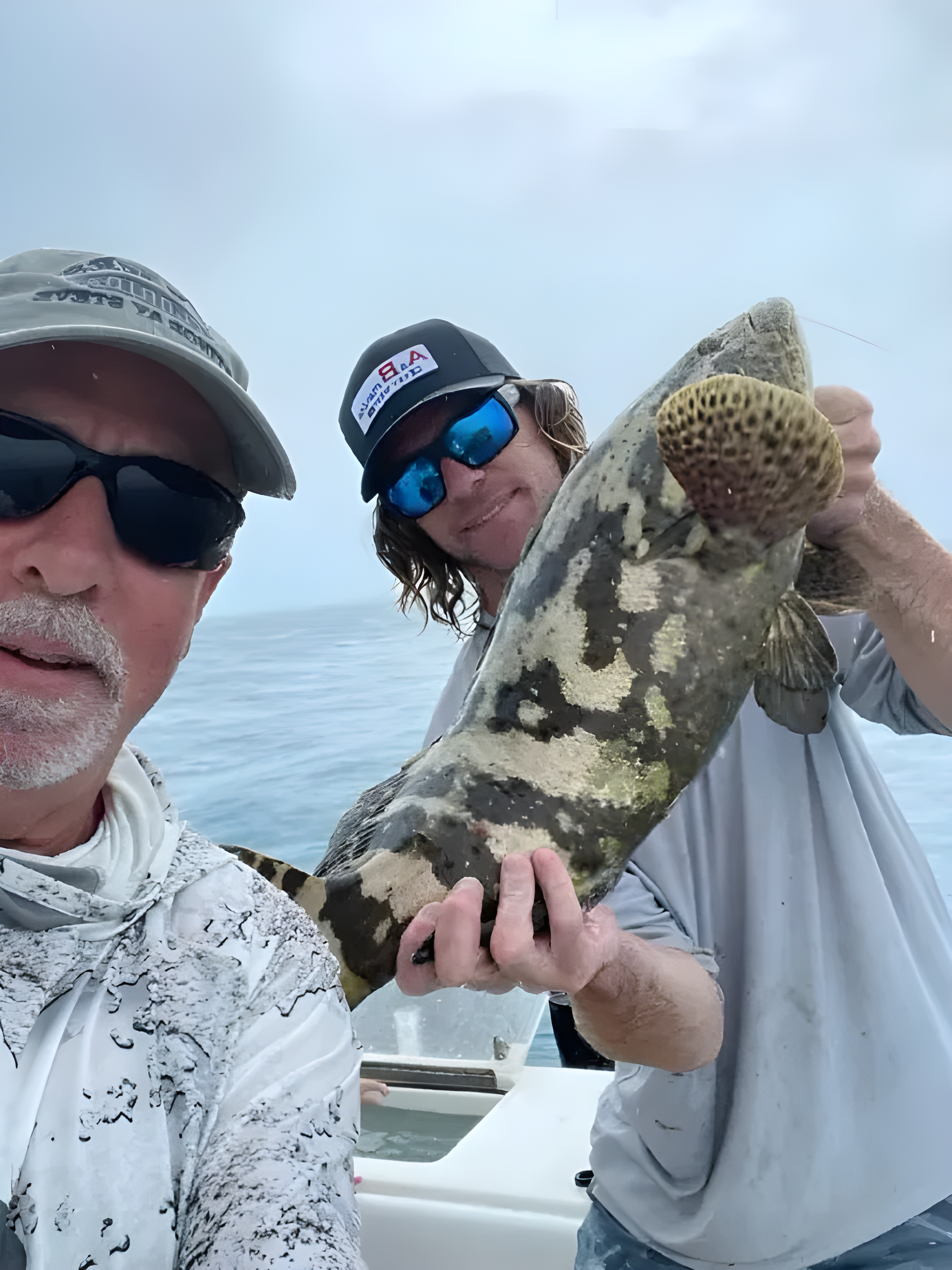 Two men on a boat holding up a large mottled fish over the water.