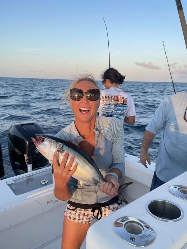 Woman smiling on a boat, holding a large fish at sea with two people in the background.