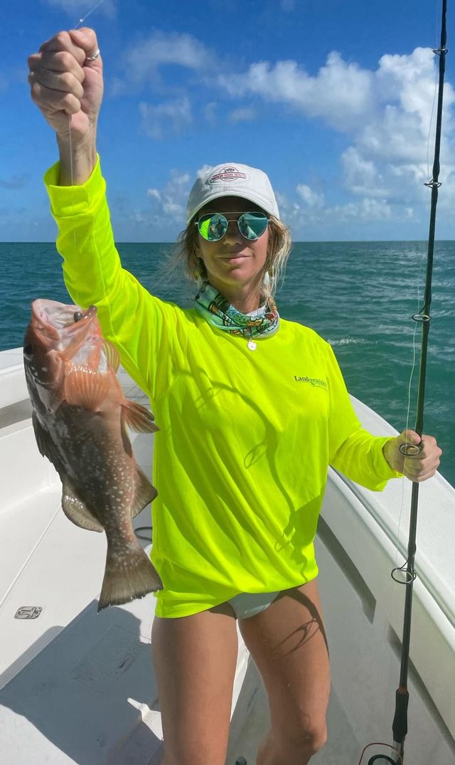 Person on a boat holding up a large fish, wearing neon green shirt and cap on a sunny day.