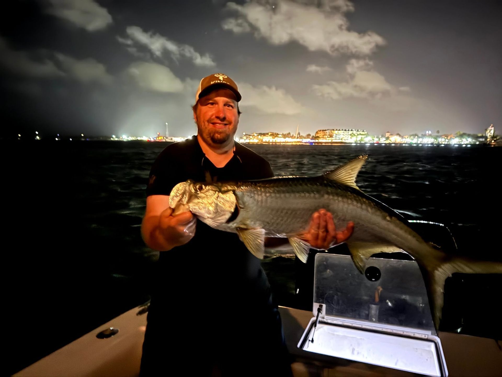 Man on a boat at night holding a large fish, with city lights glowing across the water.