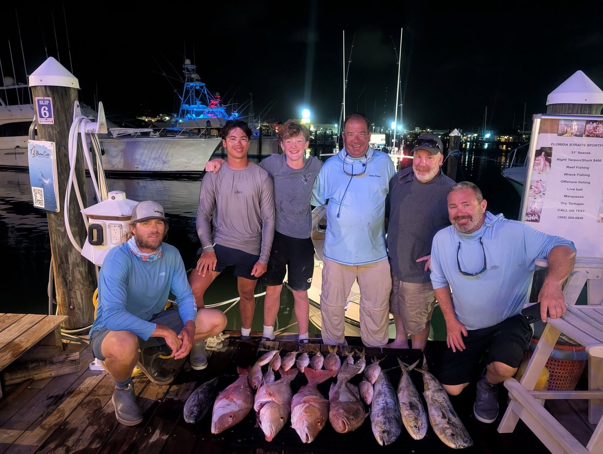 Six people posing with a large catch of fish on a dock at night, with boats and lights in the background