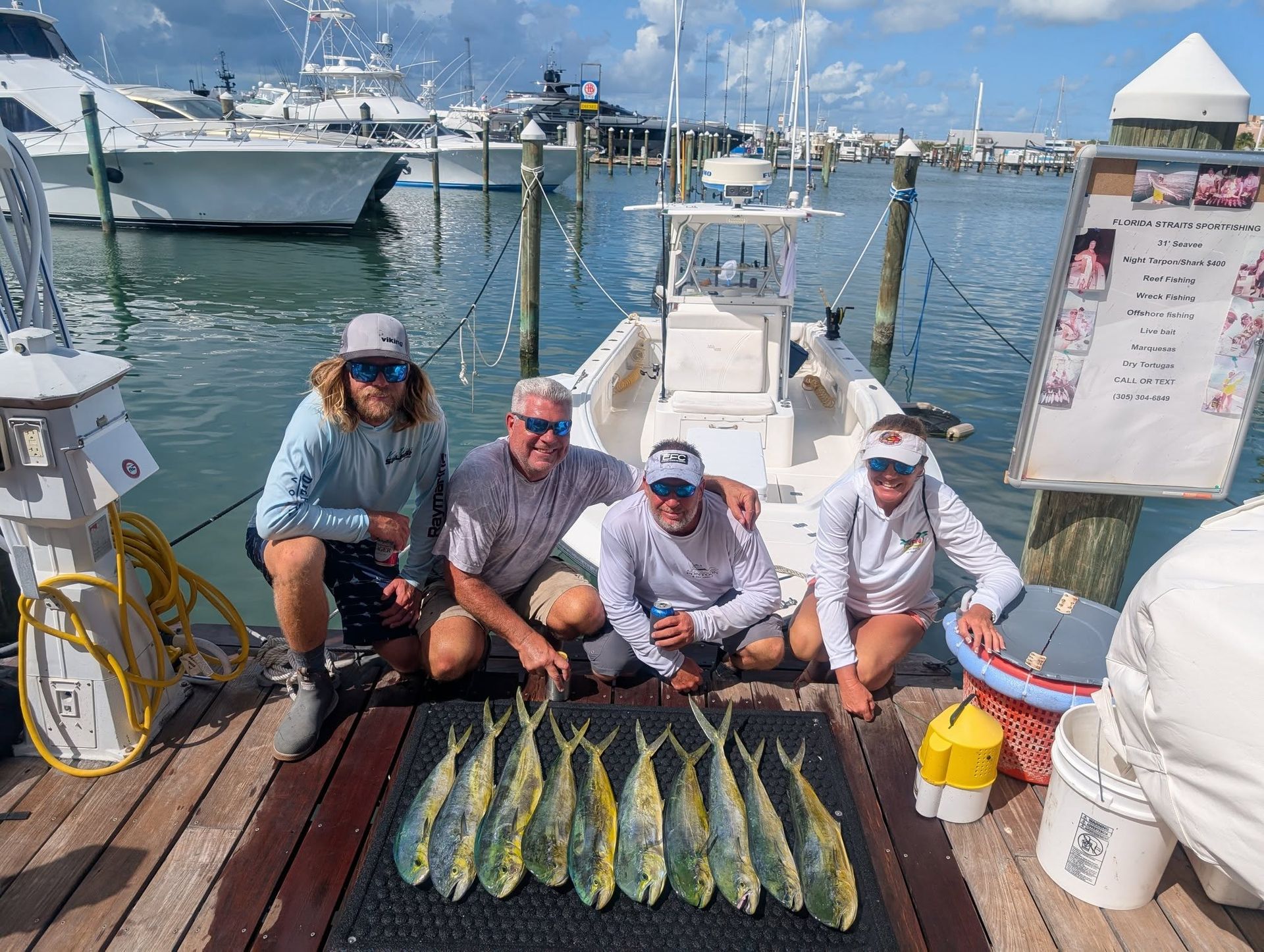 Four people on a dock pose with a row of freshly caught fish, marina boats in the background