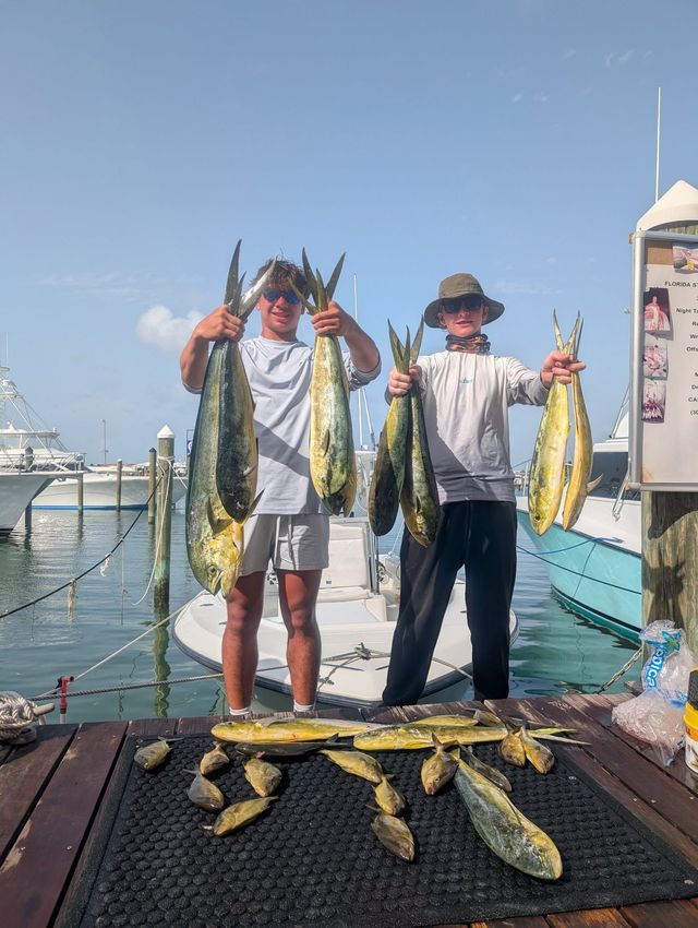 Two fishermen holding large fish on a dock beside water, with boats and a blue sky in the background.