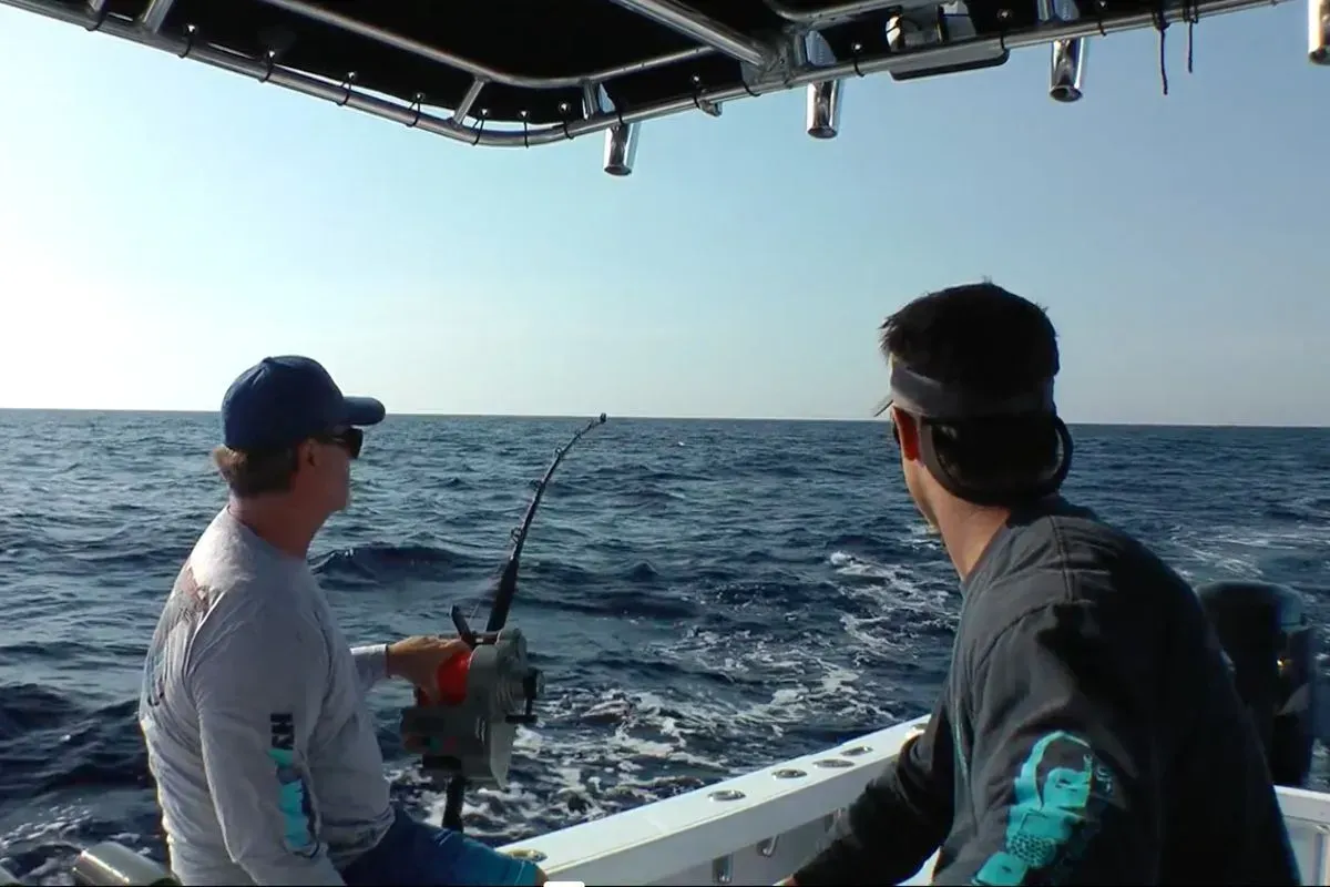Two people fishing from a boat on open water, with rods out and another boat visible on the horizon.