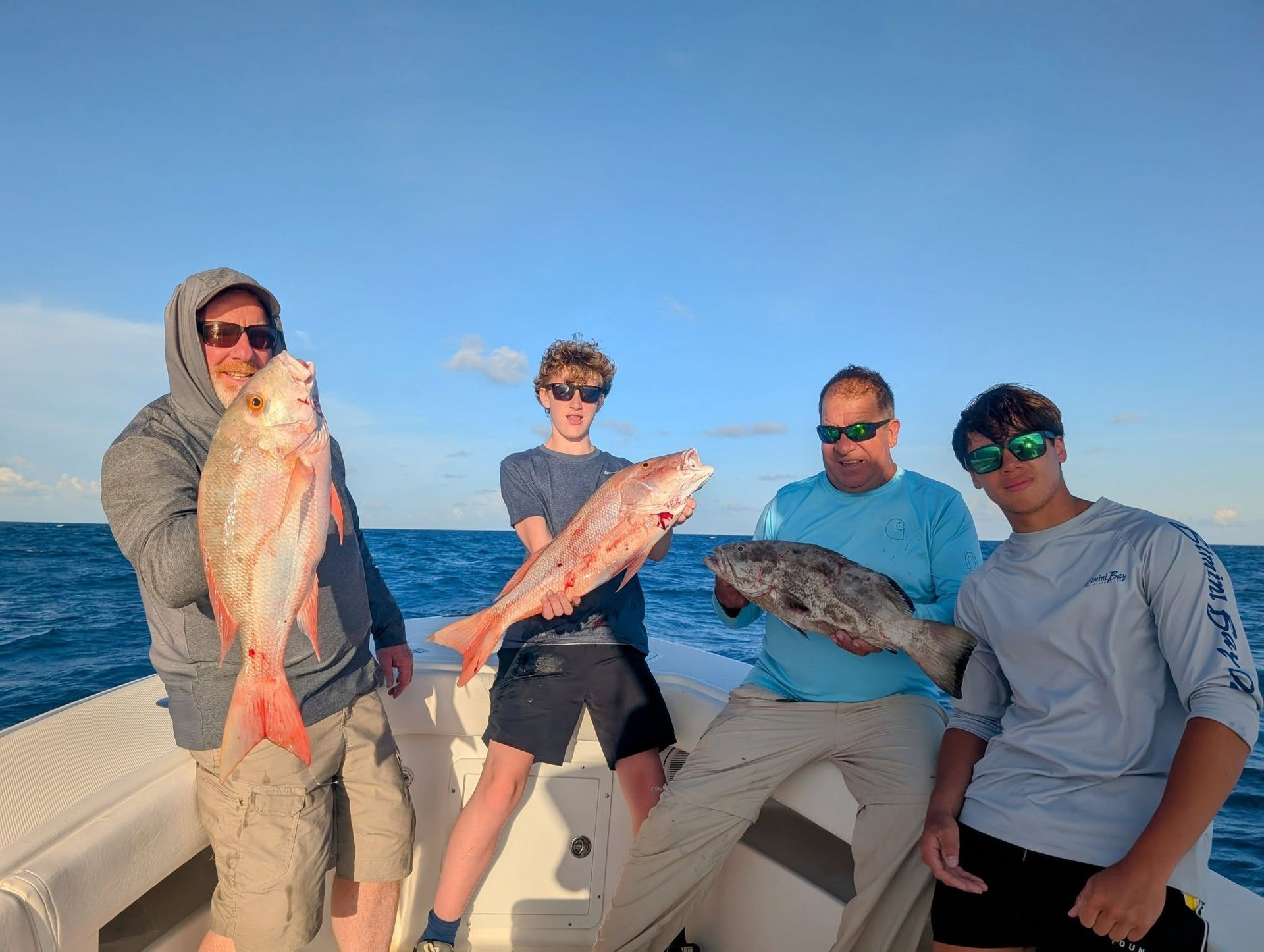 Four people on a boat holding large fish against a bright blue ocean and sky.