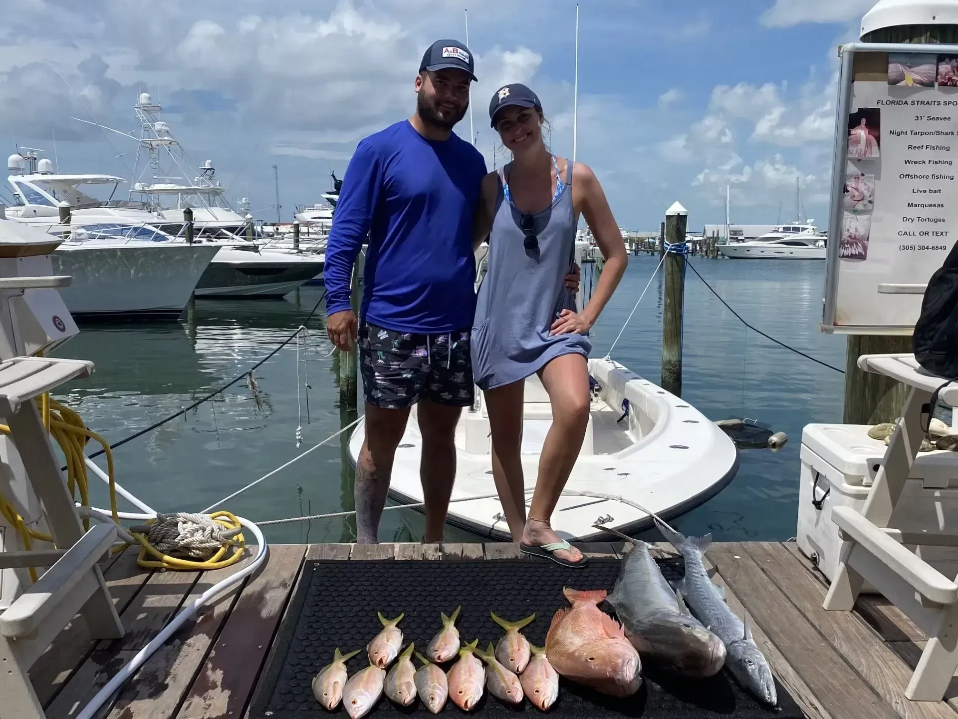 Couple on a dock with several fish laid out beside them, yachts in the marina behind them.