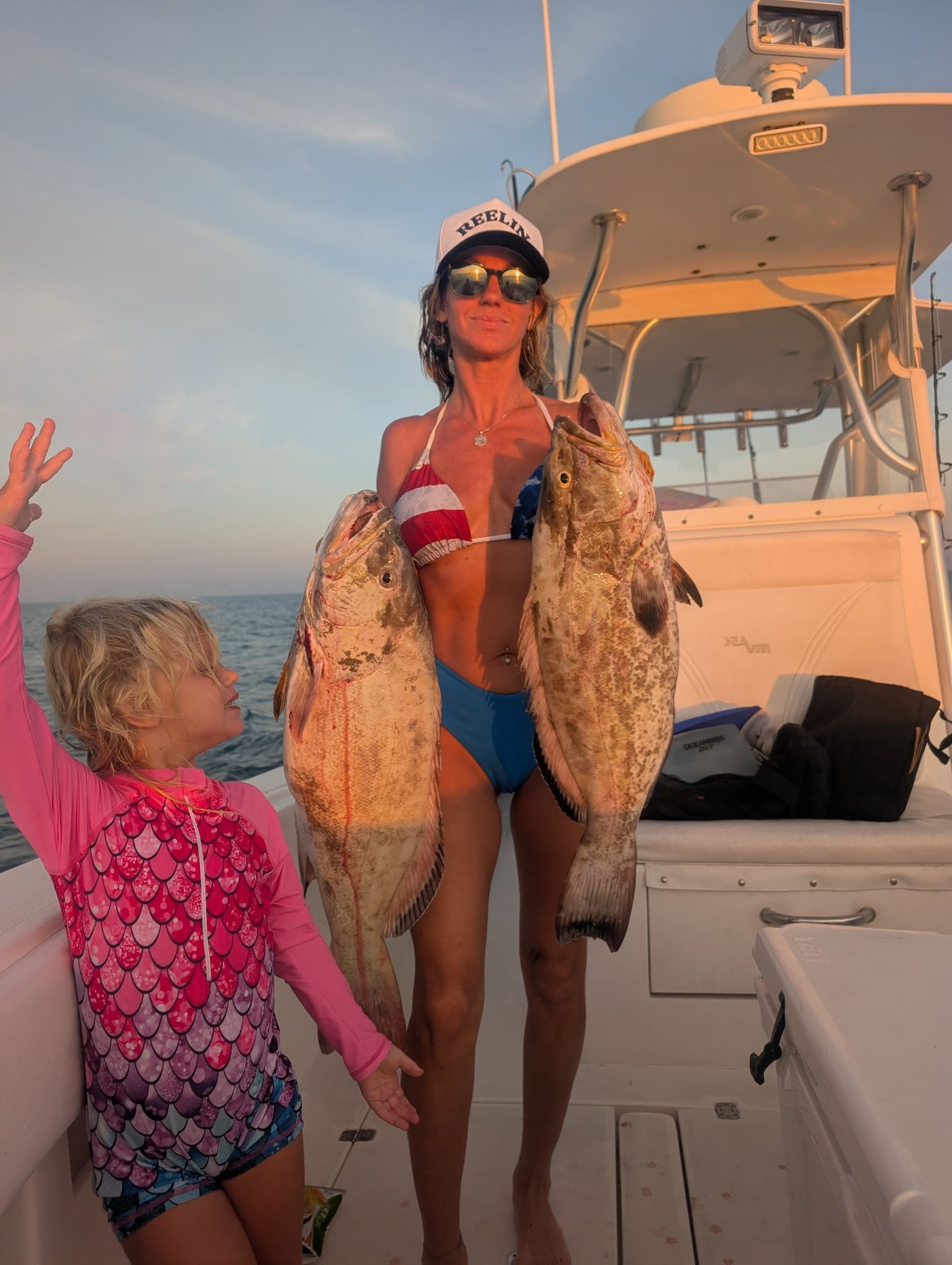 Person on a boat holding two large fish, with a child nearby at sunset.