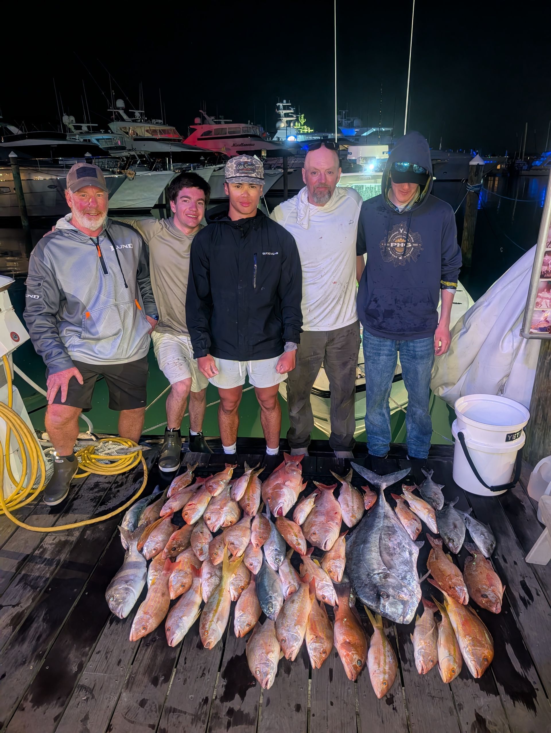 Four people on a dock at night posing behind a large catch of fish laid out on the ground