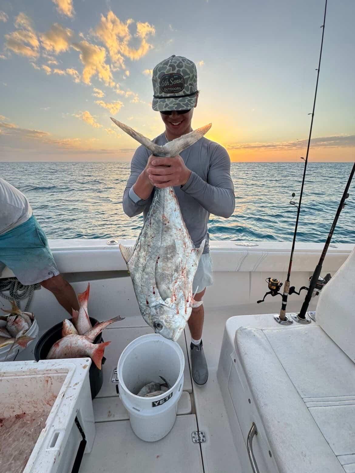 Person on a boat holding a large silver fish at sunset, with fishing rods and ocean behind.