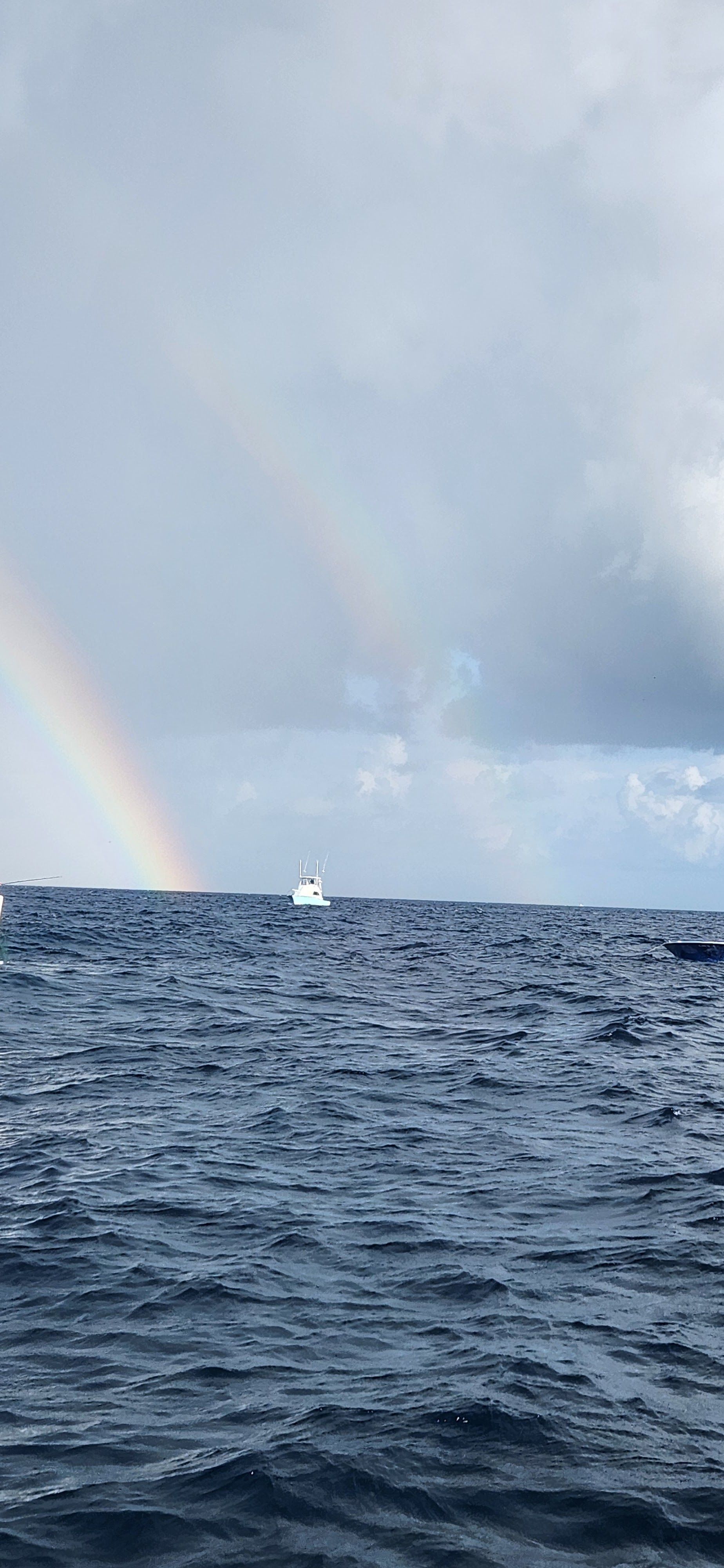 Ocean horizon under cloudy sky with a faint rainbow and a small sailboat in the distance