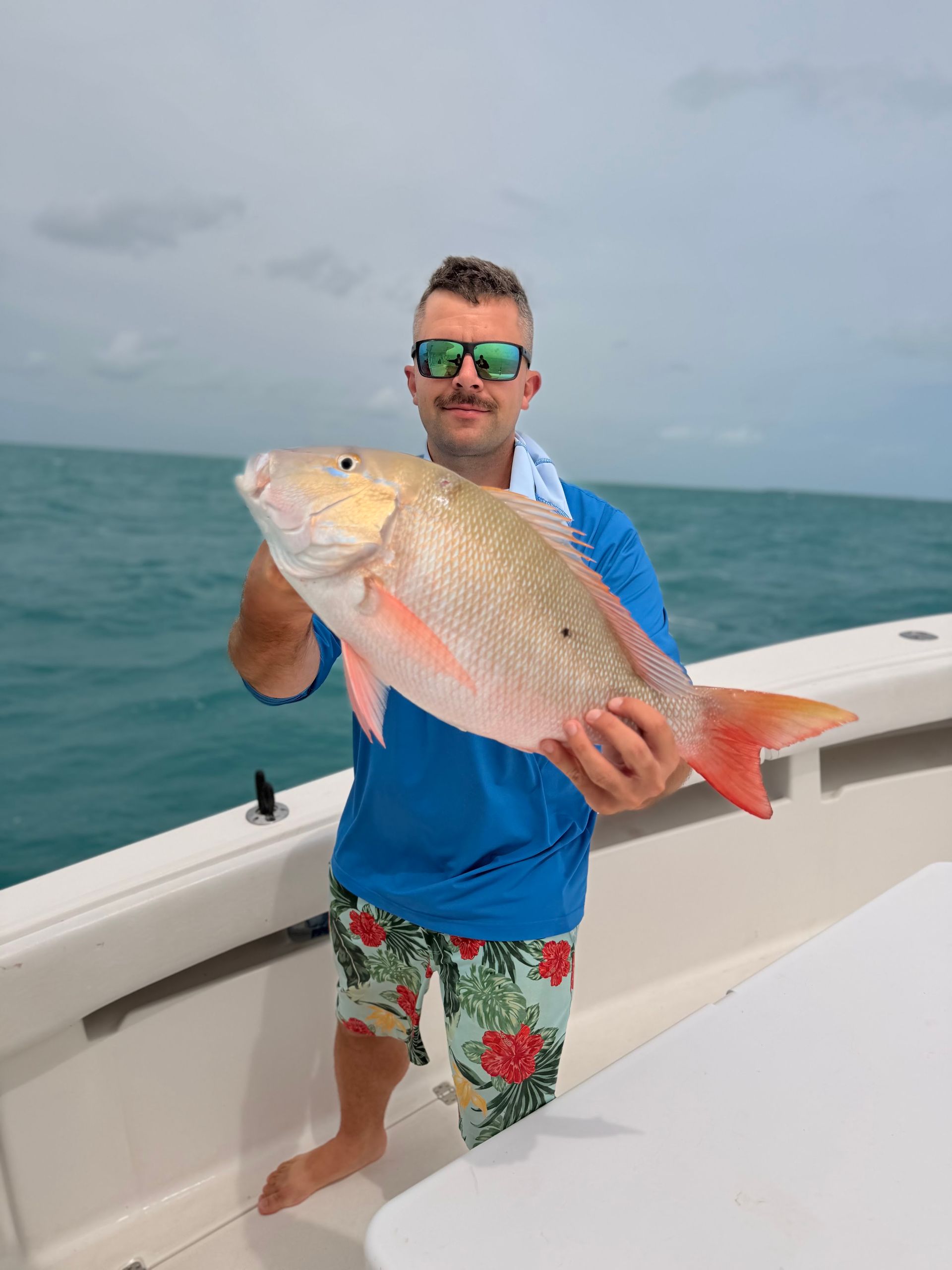 Man on a boat holding a large pink fish, wearing a blue shirt and floral shorts on the ocean.