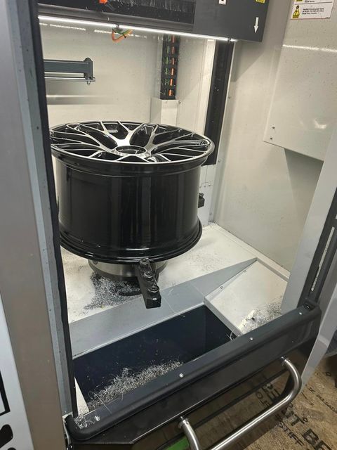 A black car wheel being machined inside a CNC machine, metallic shavings visible.