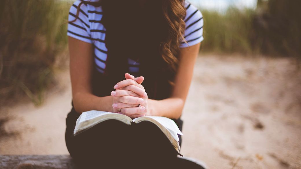 A woman is sitting on a bench with her hands folded and reading a bible.
