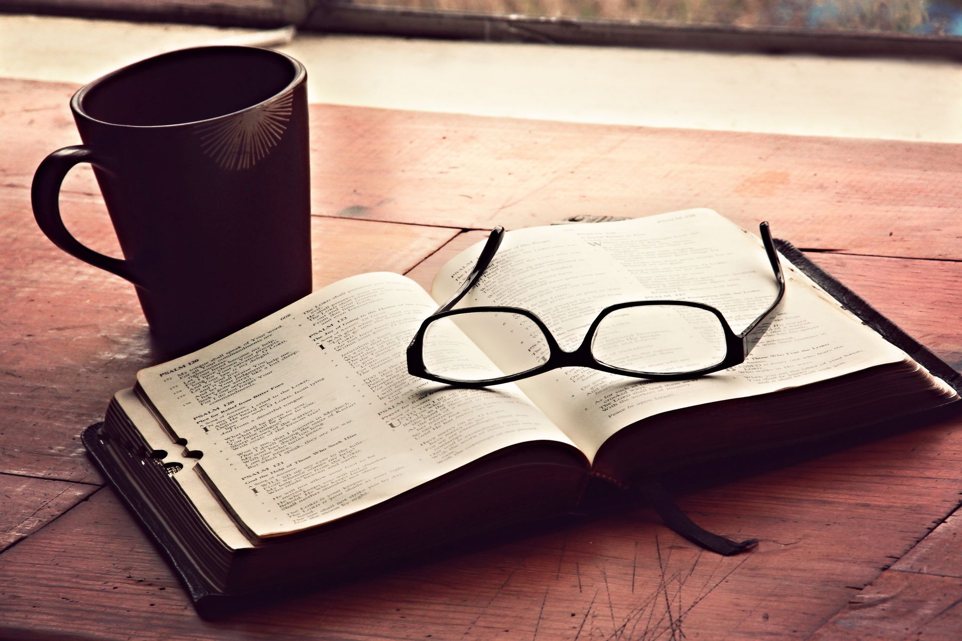 A pair of glasses sits on top of an open book next to a cup of coffee