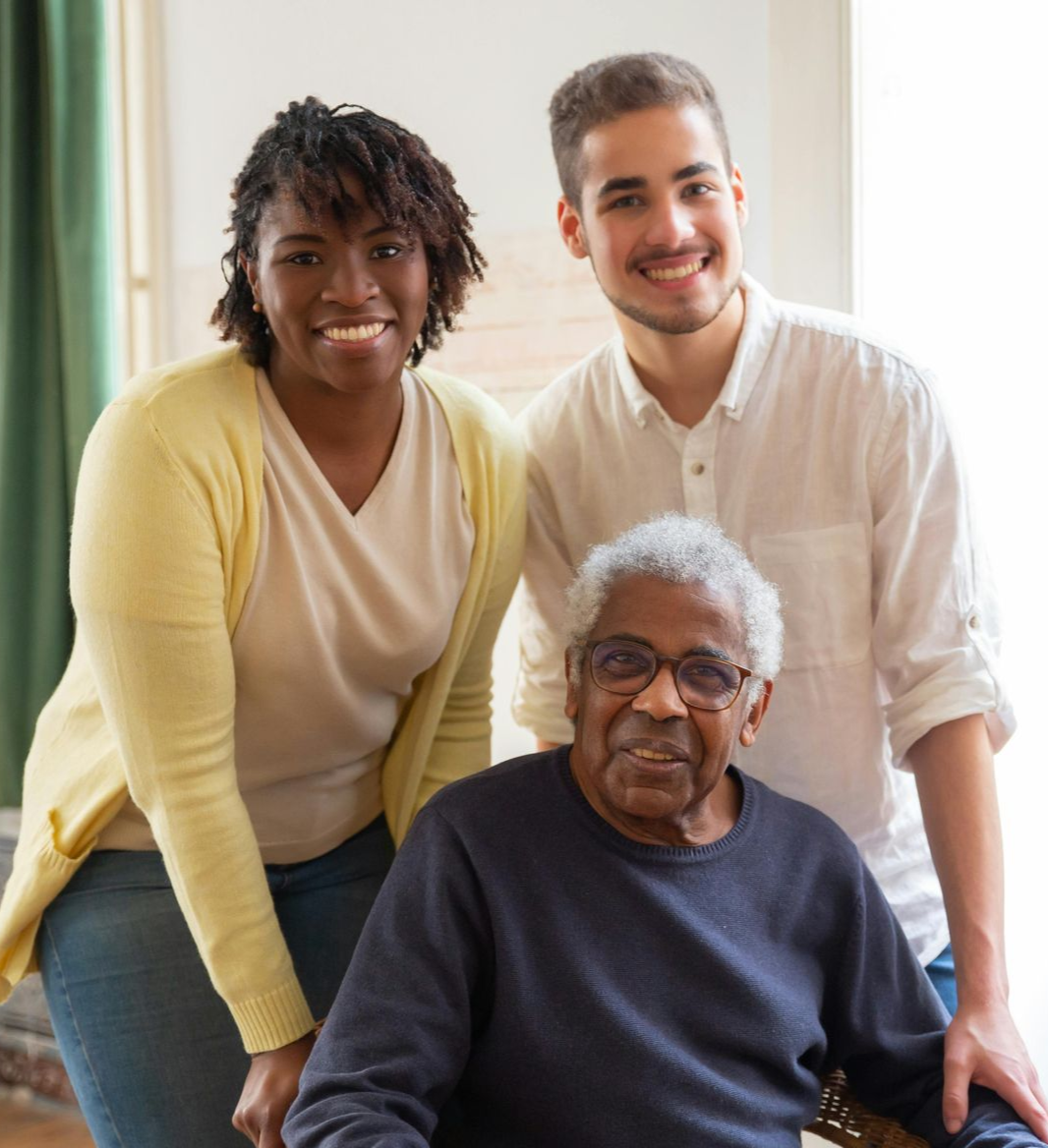Two young adults smiling with an elder in a wheelchair indoors.