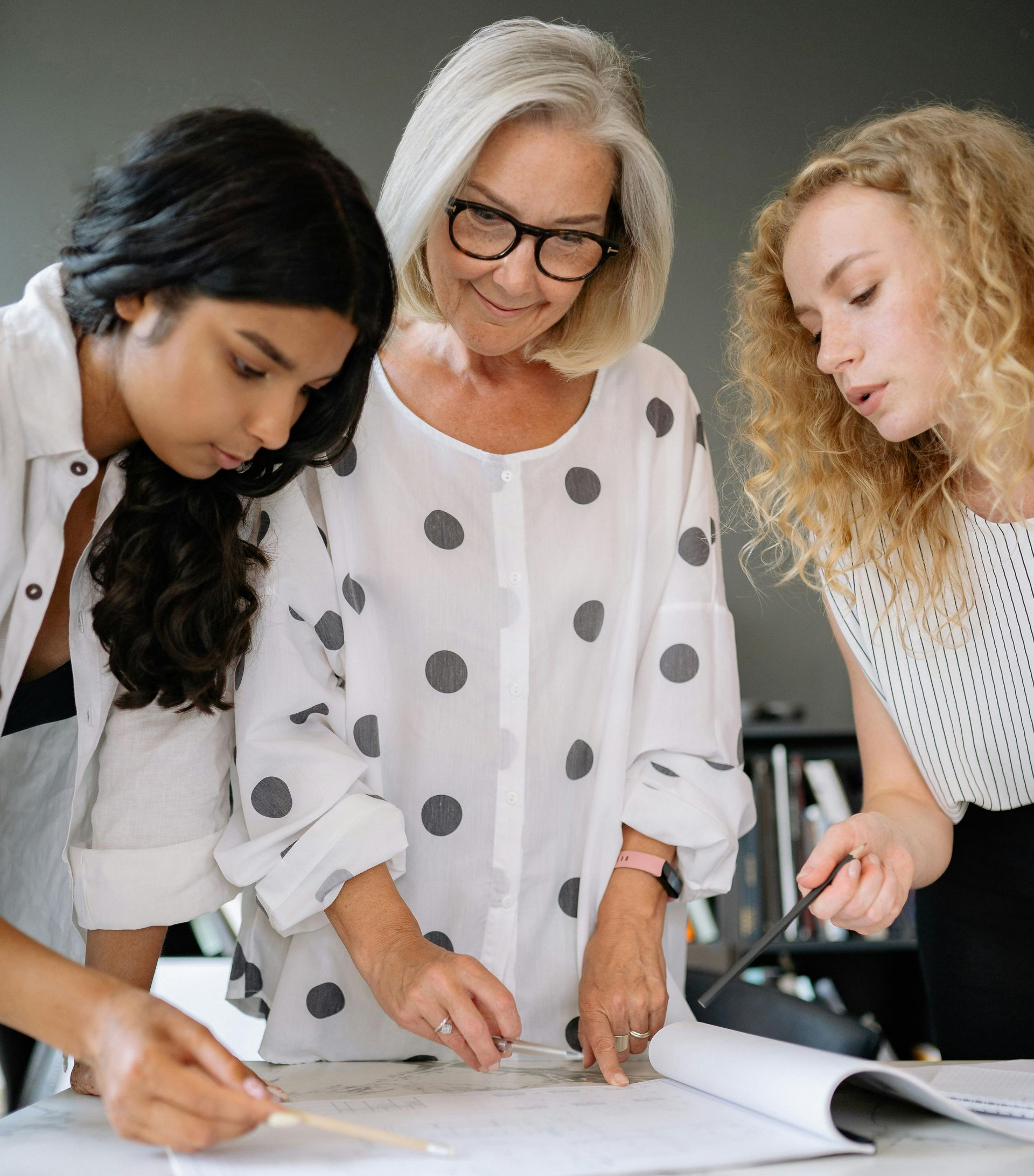 Three people collaborate around a desk, looking down at a document while discussing a project.