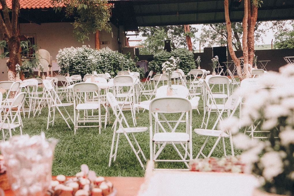 Outdoor wedding setup with white folding chairs on grass under a canopy, surrounded by flowers