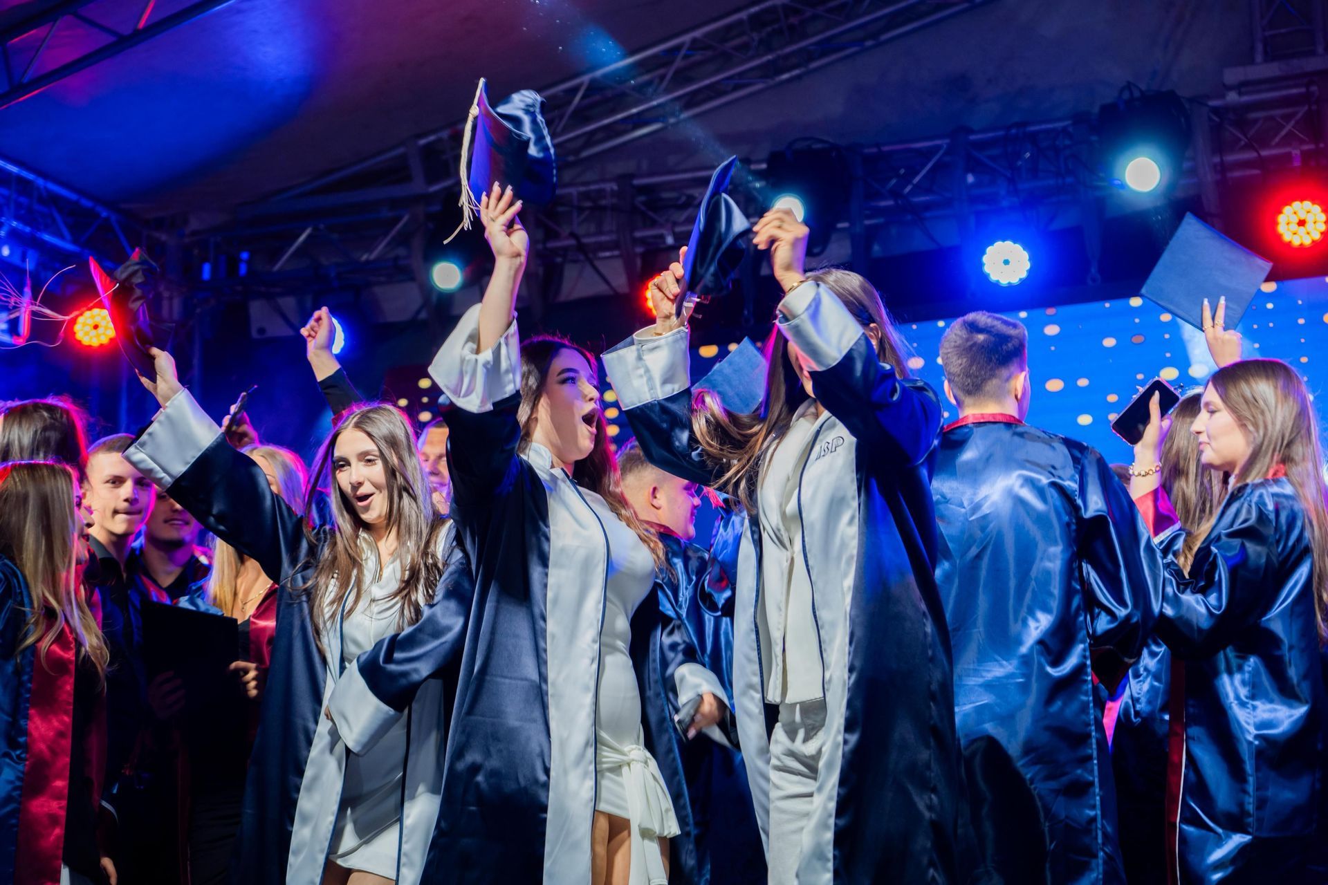 People cheering and dancing onstage under colorful concert lights, hands raised with phones.