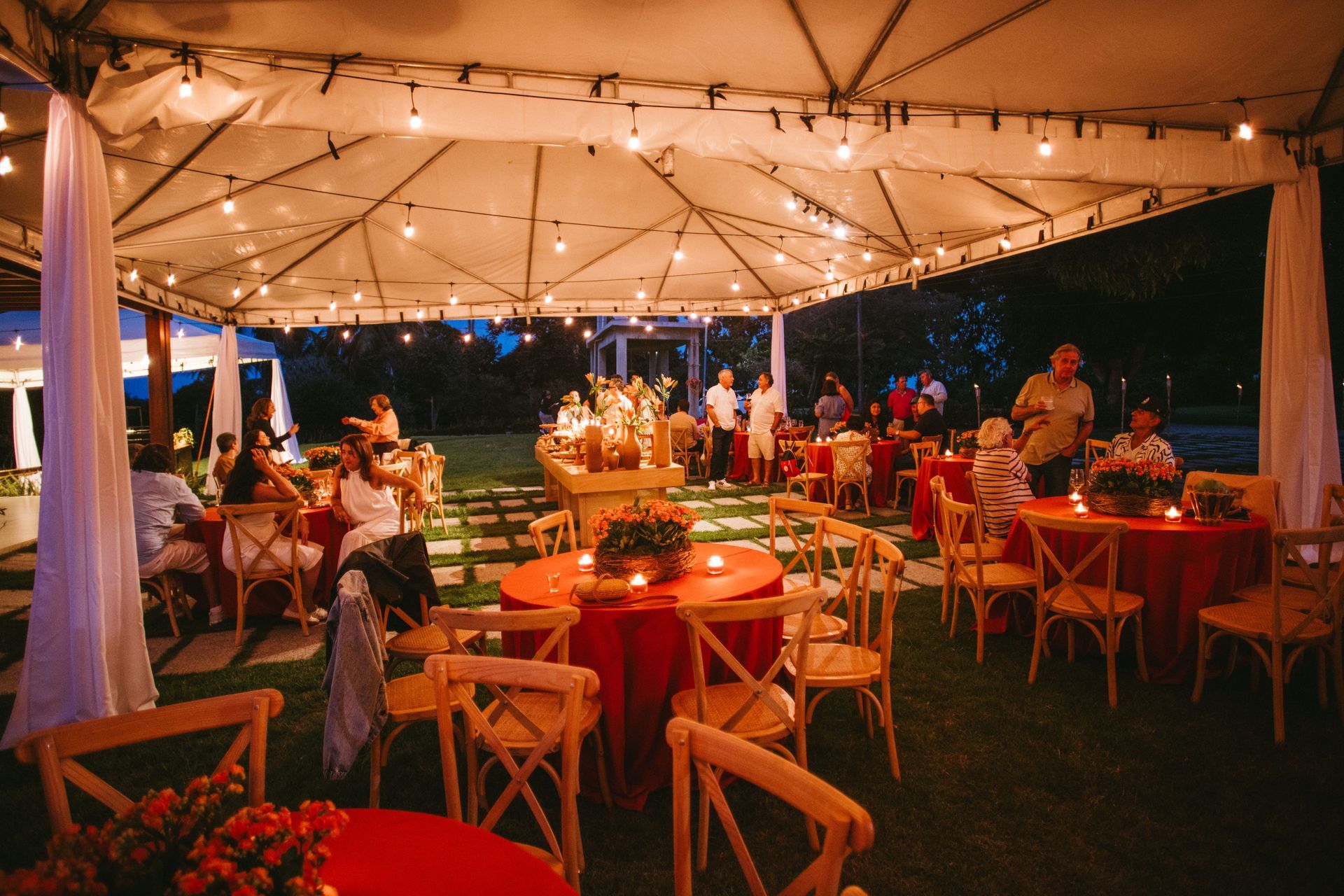 Warmly lit outdoor tent dinner with guests seated at tables and string lights overhead