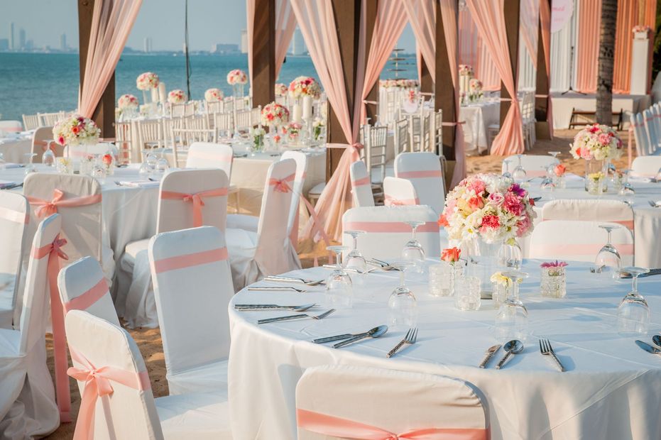 Elegant seaside wedding reception with white tables, pink chairs, and floral centerpieces under draped canopies