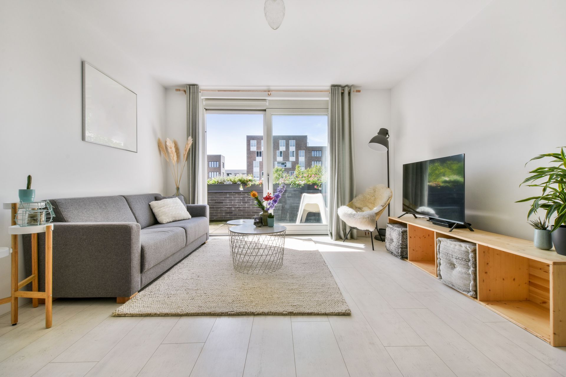 Bright modern living room with gray sofa, TV, plants, and balcony doors overlooking a city view