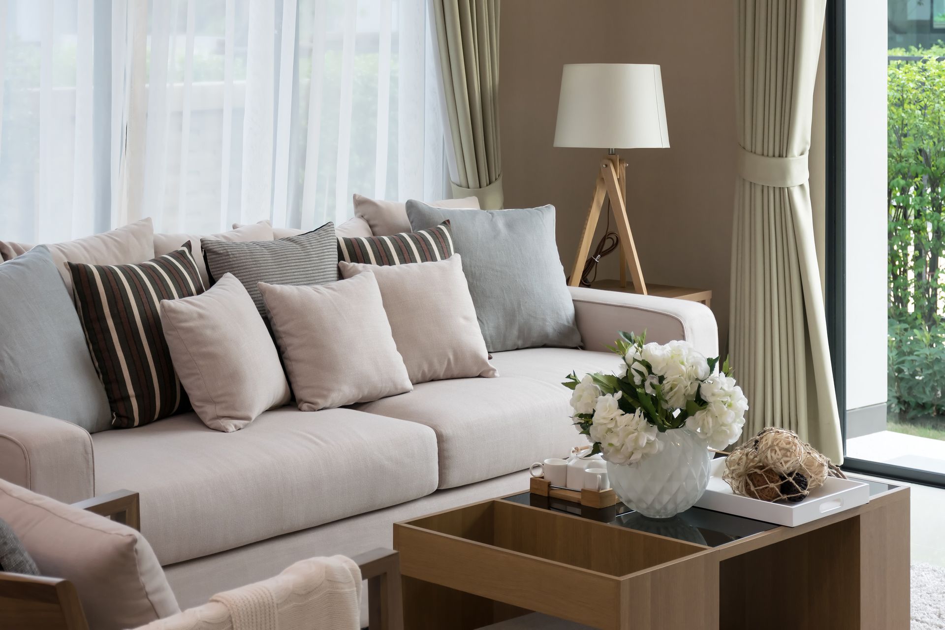Bright living room with beige sectional sofa, patterned pillows, coffee table, lamp, and large windows with curtains
