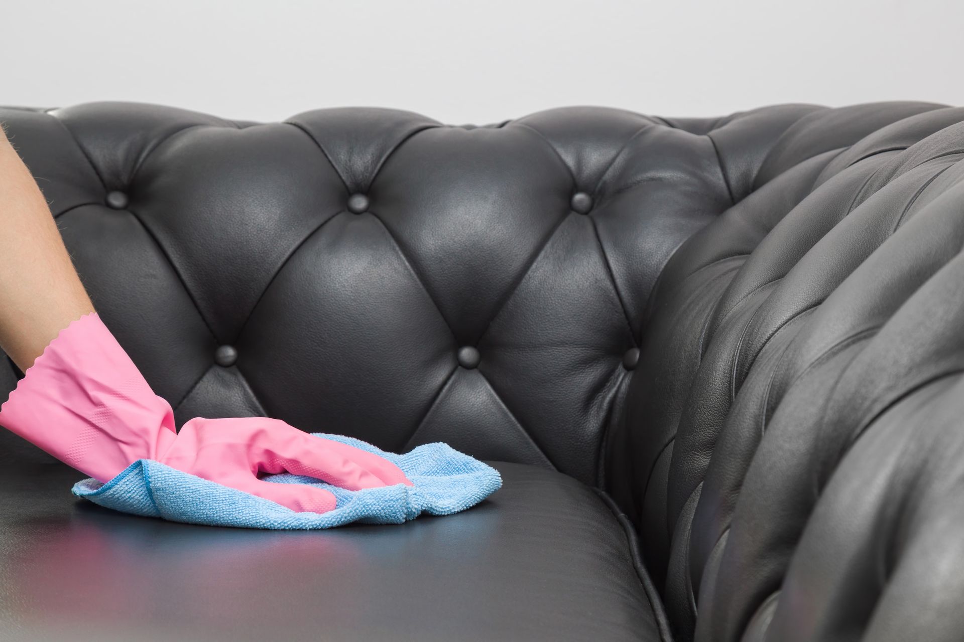 Pink-gloved hands cleaning a black leather sofa with a blue cloth.