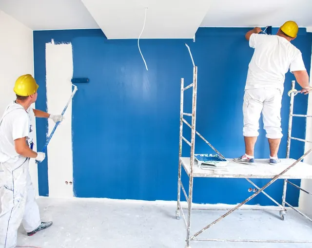 Two Painters in White Overalls and Yellow Hard Hats Painting a Blue Wall — MDSR Group In Manunda, QLD
