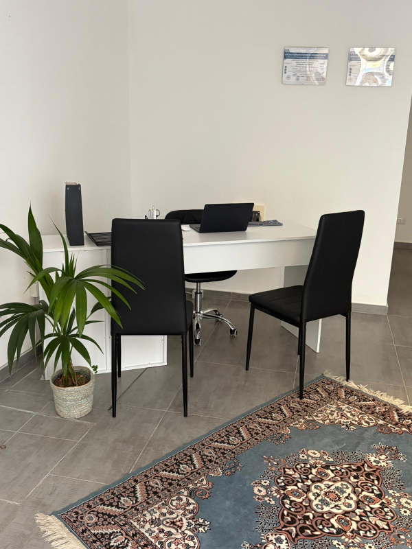 Piccola zona pranzo in ufficio con tavolo bianco, sedie nere, computer portatile, pianta in vaso e tappeto a fantasia.