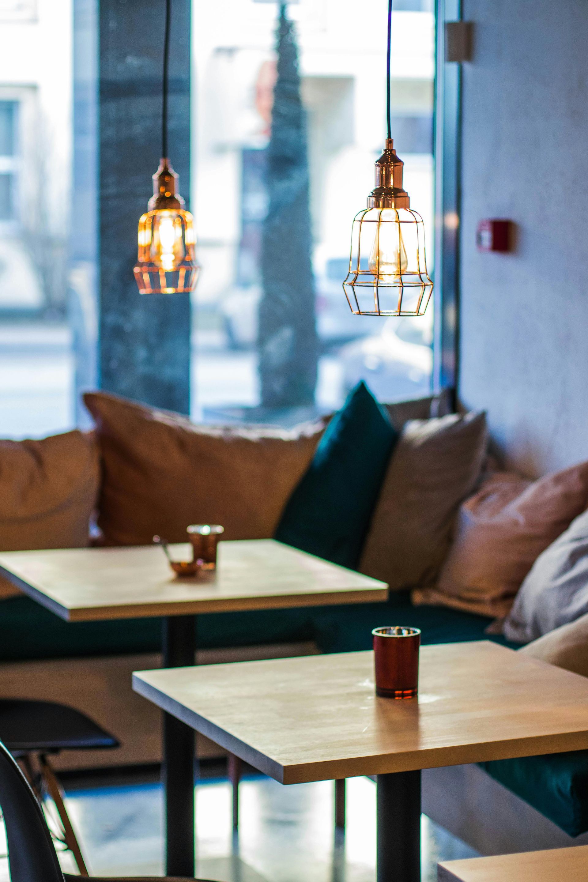 Cafe seating area with two small wooden tables and cushion seating against a large window; copper pendant lights hang above.