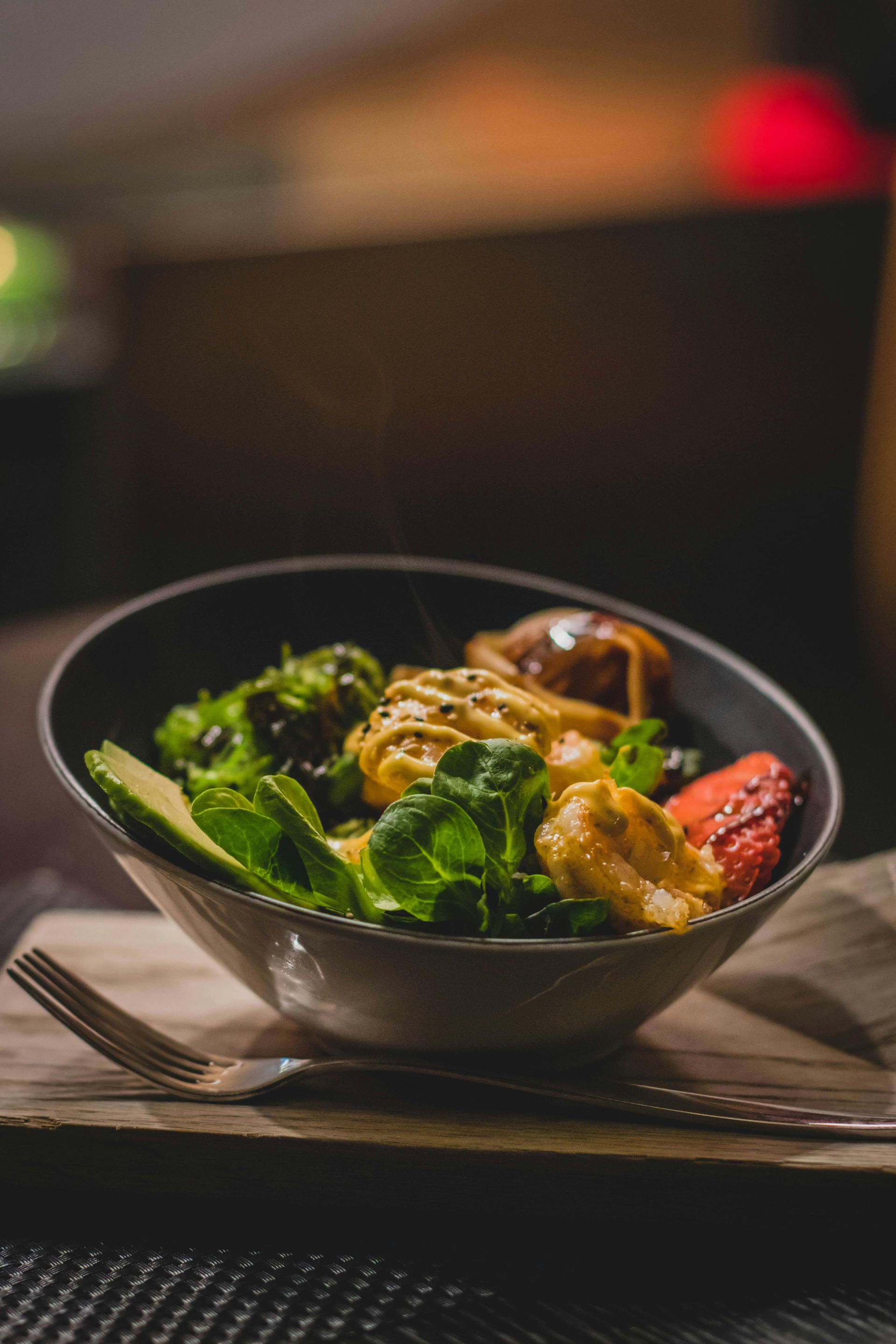 Bowl of salad with various greens, avocado, and a sauce, set on a wooden board with a fork.