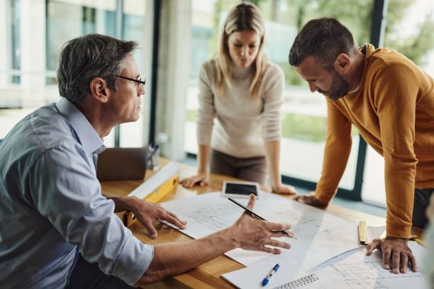 Three people review documents and discuss plans around a table in a bright office.
