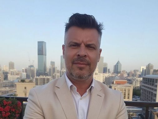 Man in a beige blazer stands on a rooftop with a city skyline in the background.