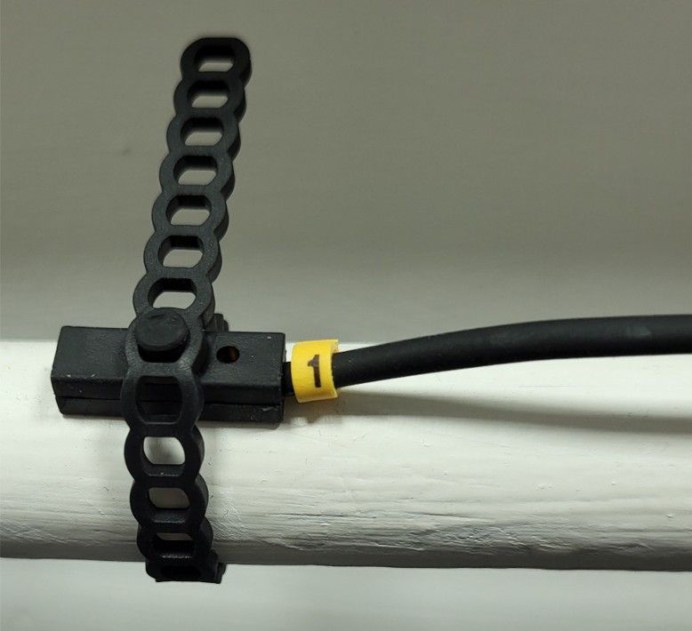 Black cable clipped to a white pipe with a black strap and a small yellow tag labeled “1”.