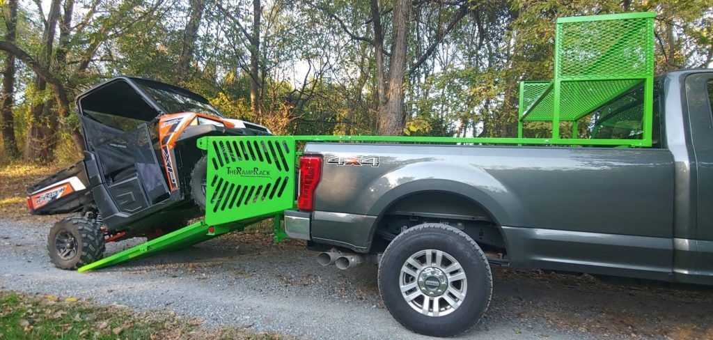 Green loader ramp on a gray pickup, loading a black-and-orange machine on a gravel road in the woods