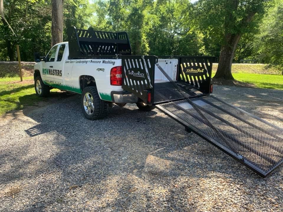 White pickup truck with extended metal ramps parked on gravel in a wooded area.