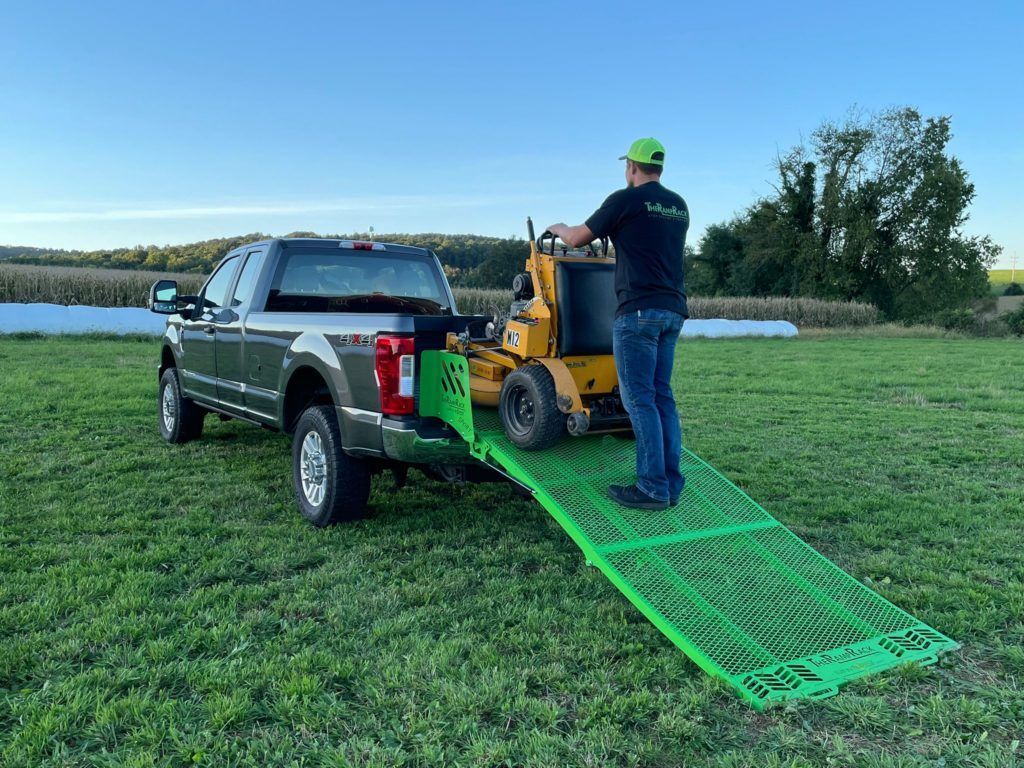 Man in green helmet loading yellow machine into pickup using a green ramp in a grassy field