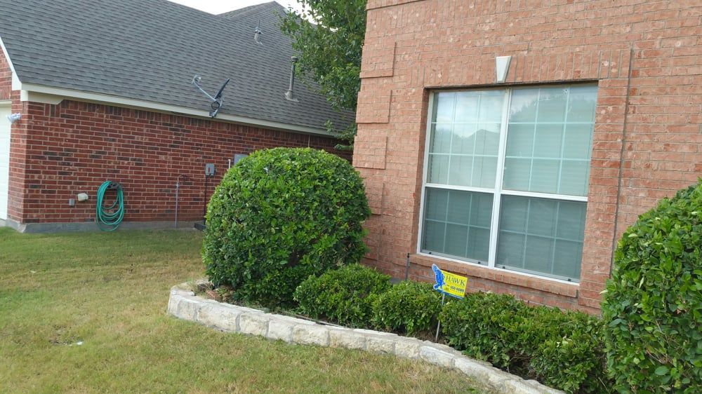 Brick house corner with a large window, trimmed bushes, and a curved stone garden border