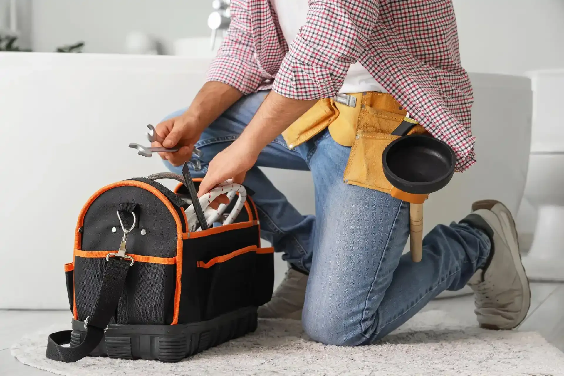 Idraulico inginocchiato, che seleziona gli attrezzi da una borsa portautensili nera e arancione in un bagno.