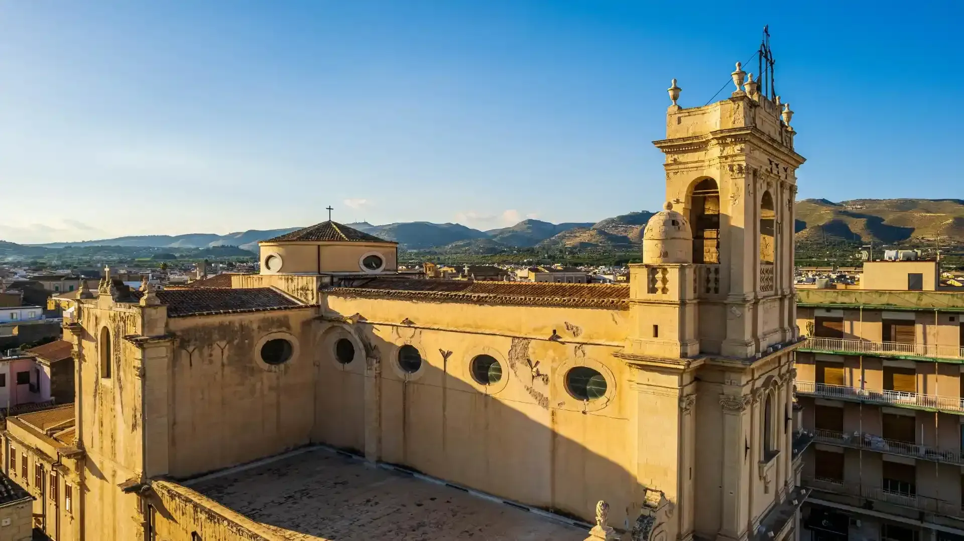 Vista esterna di un'alta cattedrale dai colori chiari, con un campanile sotto un cielo azzurro e il paesaggio urbano sullo sfondo.