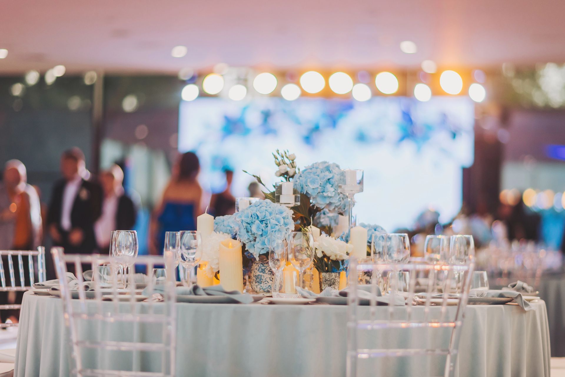 Tavolo rotondo apparecchiato per l'evento, decorato con fiori blu, candele e sedie trasparenti. Persone sullo sfondo.