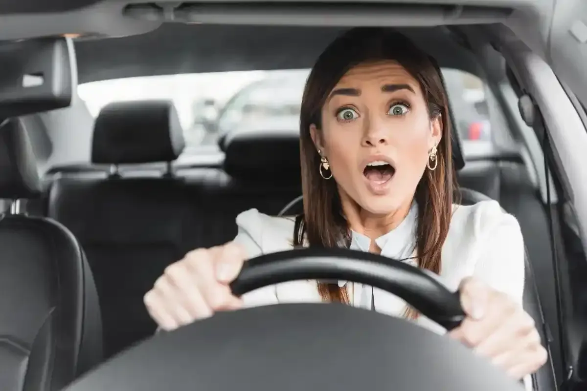 Donna con gli occhi spalancati, che stringe il volante dell'auto; sembra sorpresa.