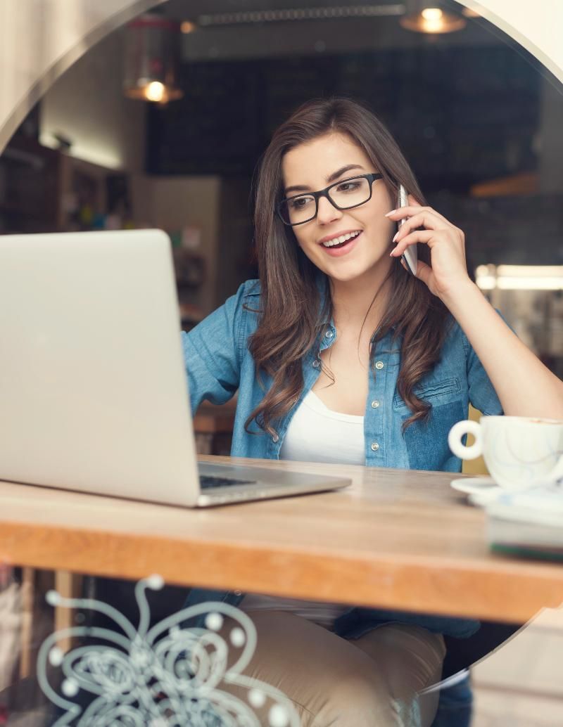 Donna al bar, che usa il computer portatile e il telefono, sorridente, con una tazza di caffè.