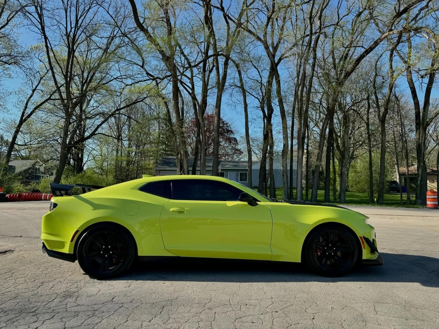 Bright yellow sports car with black wheels parked on pavement, trees in the background.