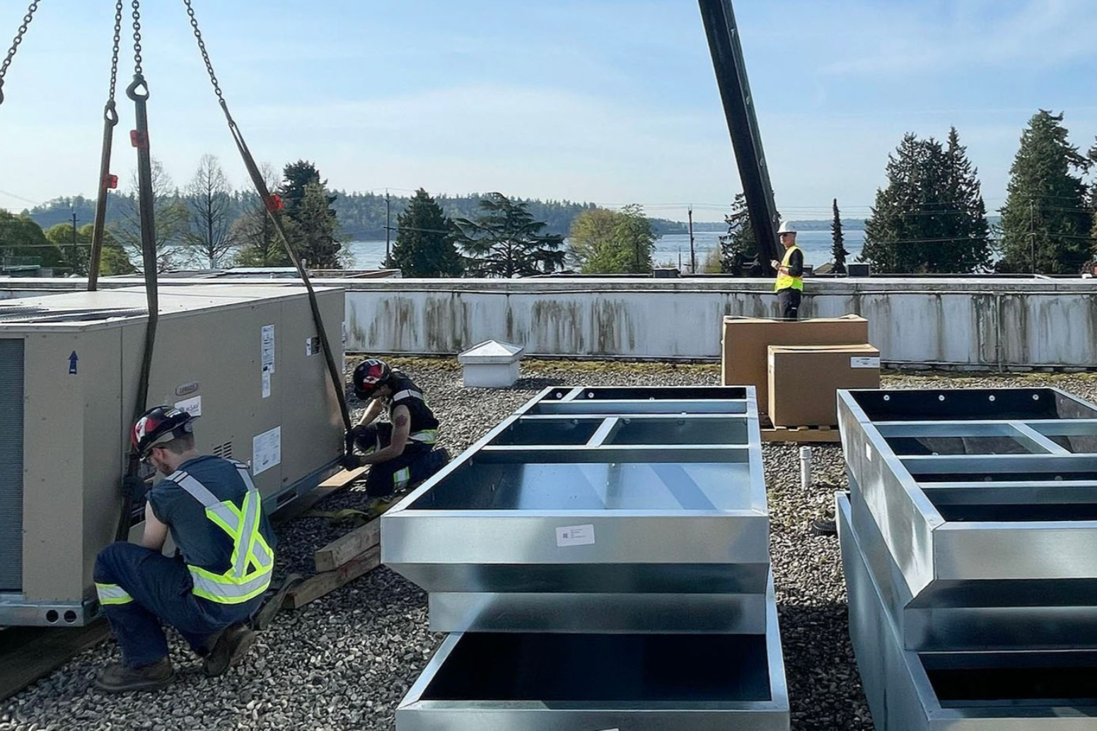 Workers installing HVAC unit on a rooftop with crane. Blue sky, lake in background.