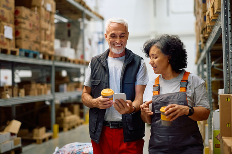 Two people in a warehouse looking at a phone, smiling and holding coffee. Shelves of boxes are in the background.
