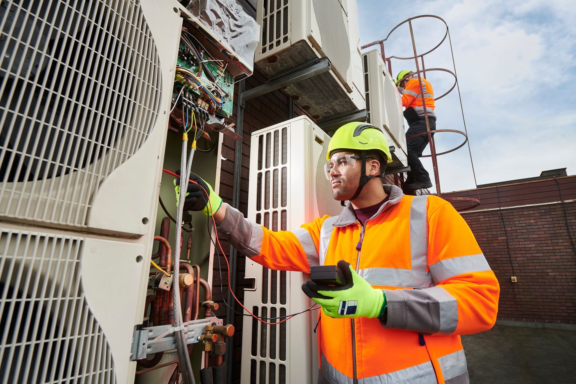 Two HVAC technicians in safety vests working on air conditioning units outdoors.