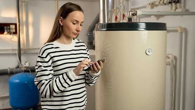 Plumber in blue uniform, holding wrench, smiling in front of water heater and pipes.