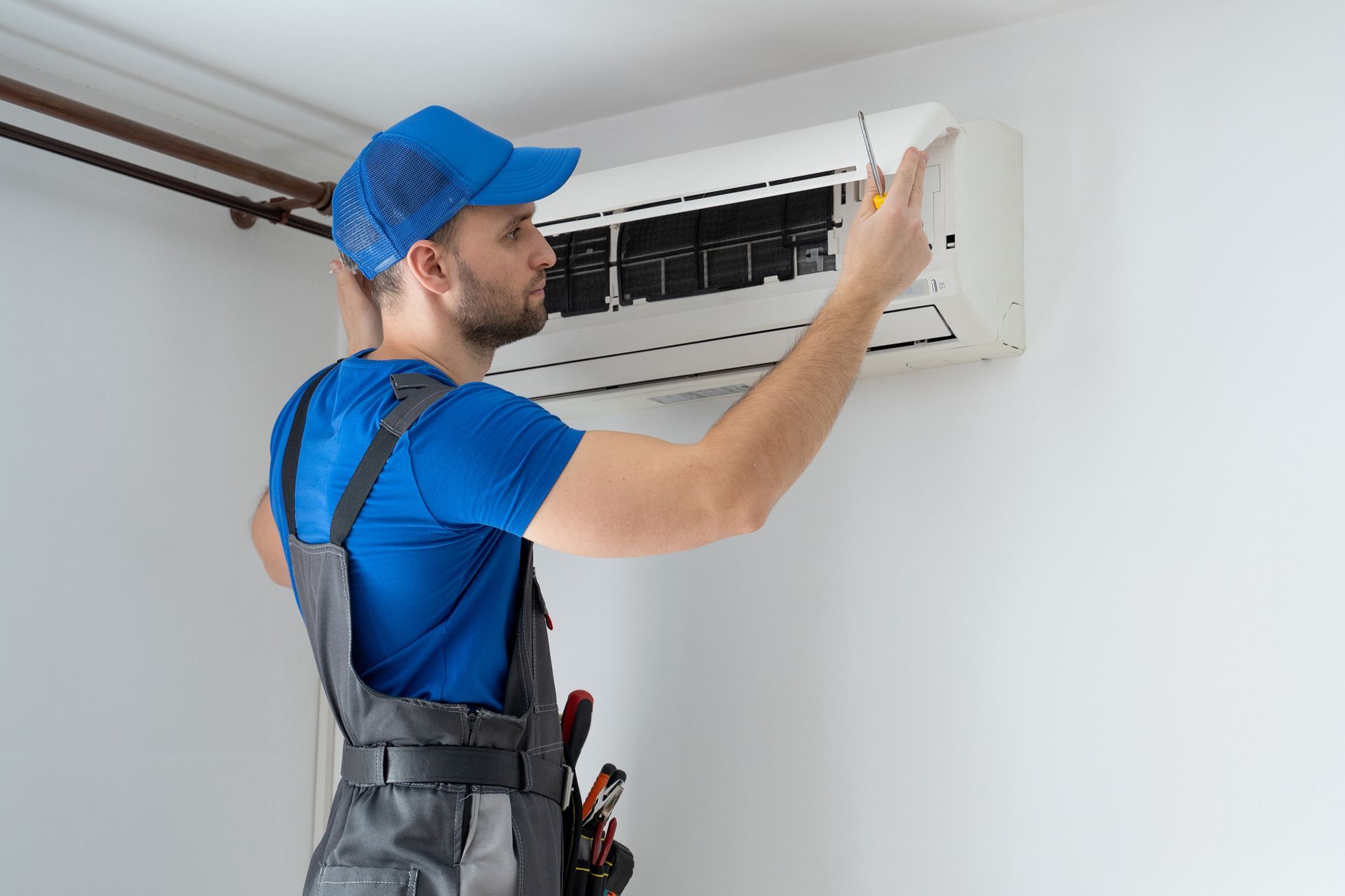 Technician in blue uniform repairing a wall-mounted air conditioner with tools in a white room.